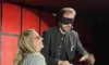 A woman laughing on stage stands next to a blindfolded man holding a folded newspaper.