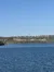 A serene lake stretches across the foreground with a hilly landscape featuring scattered buildings and a water tower under a clear blue sky.