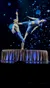 Two acrobats in blue outfits perform a balancing act on a circular platform against a backdrop of floating blue lights.