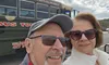 A couple wearing sunglasses takes a selfie in front of a green tour bus with the phrase