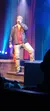 A person in a black jacket and distressed jeans is singing on stage under blue and purple lighting.