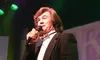 A man in a black tuxedo with decorative side seam details is performing on stage while holding a microphone.