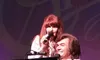 A woman is singing into a microphone while a man plays a keyboard, both appearing to perform on stage.