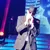 A performer in a black suit, hat, and sunglasses is singing into a microphone on stage.