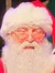 A person dressed as Santa Claus stands on a festive stage, holding a large red sack in front of a green Christmas tree backdrop.
