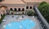 The image shows an aerial view of a uniquely shaped swimming pool and surrounding lounge area beside a large building with a red roof, set amidst a backdrop of lush greenery.
