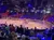 A crowd watches a rodeo performance featuring riders on horses carrying American flags in a brightly lit indoor arena.