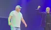 Two men are on stage, one wearing a striped shirt and cap, while the other raises his fist, under blue and green lighting.