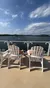 Two people are relaxing in Adirondack chairs on a deck overlooking a scenic lake with a partly cloudy sky.