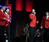 A man in a red jacket and a woman in a sparkly dress sit across from each other at a table on stage, with the man holding a microphone and a 