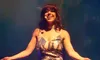 A woman is on stage wearing a sparkly gown and a headdress, with her arms open wide.