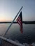 An American flag is flying on a pole over a body of water at dusk, with a scenic view of distant hills.