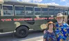 A couple wearing colorful clothing and hats stand smiling in front of a bus labeled