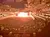 The image depicts an indoor arena with a sand floor, where spectators are watching a bright explosion or special effect near the stage area.