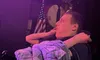 A person in a plaid shirt is sitting in a dark room, looking up at a colorful, illuminated screen, with a US flag and a large drum in the background.