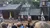 A group of people on horseback hold flags in front of a rustic wooden building, while a crowd watches.