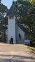 The image shows a quaint, white wooden church with a bell tower, surrounded by trees and a clear blue sky.