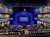 The image shows a theater interior with an audience seated, facing a stage adorned with a