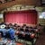 The image shows a theater setup with a stage featuring red curtains and microphones, and dining tables with seats arranged for an audience.