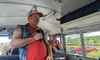 Two people are holding microphones and sitting inside a bus with several license plates on the ceiling.