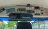 The interior of a vehicle displays various vintage license plates and humorous signs above the rearview mirror, with a view of a parking lot and buildings outside the windshield.