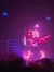 A performer in an LED suit plays guitar on a dimly lit stage with purple and blue lighting effects.