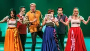 A group of performers on stage, dressed in colorful costumes, are energetically engaged in a lively act, with one holding a sign that reads