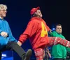 Three people are performing on stage energetically dancing and wearing colorful hoodies and festive pants