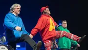 Three people are performing on stage, energetically dancing and wearing colorful hoodies and festive pants.