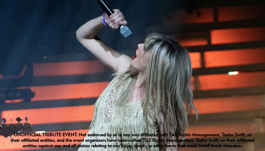 A performer in a fringed outfit is energetically singing into a microphone on stage at an unofficial tribute event.