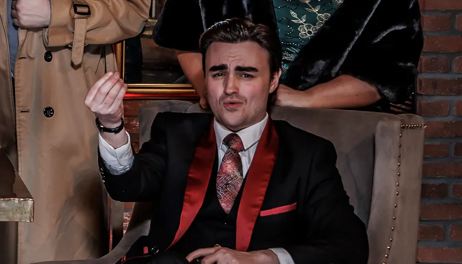 A person in a suit with a red satin collar is making a expressive gesture while sitting in a chair.
