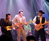 Three musicians are performing on stage with guitars under colorful stage lighting