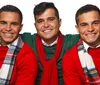 Three smiling men are posing together wearing red and green Christmas-themed clothing and colorful plaid scarves