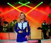 A performer in a blue and white outfit sings on a stage with a band in the background illuminated by colorful stage lights