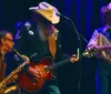 Three musicians perform on stage one playing saxophone another electric guitar and the third playing violin all wearing cowboy hats