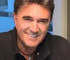 A man with short hair smiles warmly dressed in a dark shirt with a guitar visible in the background