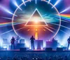 A band performs on stage under a vibrant light show with a large triangular prism and rainbow projection in the background while the audience captures the moment