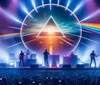 A band performs on stage under a vibrant light show with a large triangular prism and rainbow projection in the background while the audience captures the moment