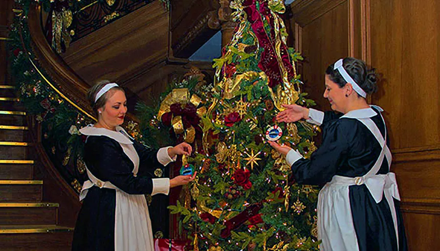 Two women in traditional maid outfits are decorating a lavishly adorned Christmas tree with ornaments near a grand staircase.
