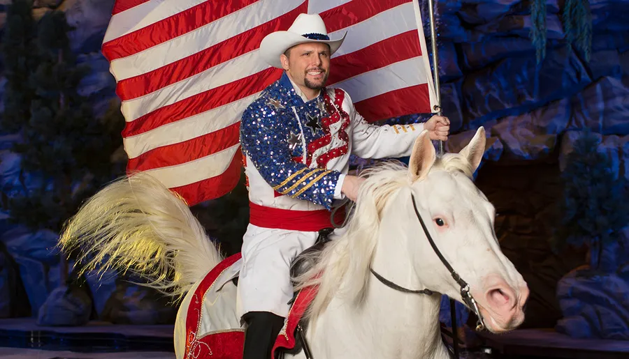 A man in a cowboy hat and a sequined patriotic outfit rides a white horse while holding an American flag.