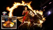A performer rides two horses through a flaming ring in a dramatic, lit-up setting.