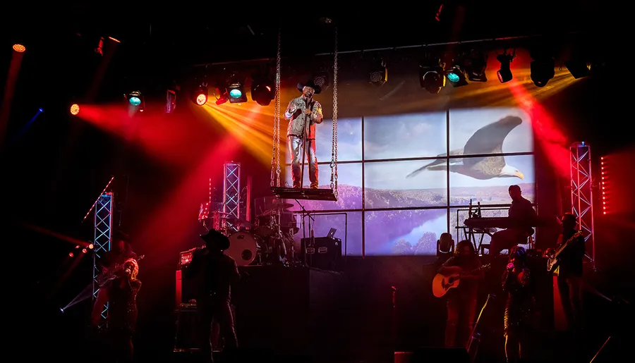 A musician performs on a raised platform among band members under colorful stage lights with an eagle displayed on the screen in the background.