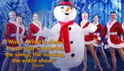 A person in a snowman costume is surrounded by smiling dancers in festive outfits on a winter-themed stage.