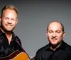 Two smiling men one holding a guitar and the other standing next to an upright bass pose for a portrait against a gray background