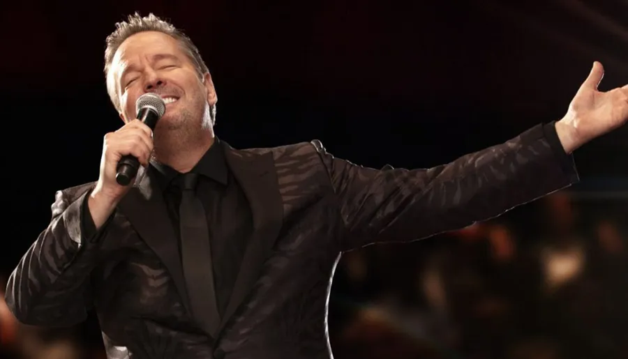 A man in a patterned suit passionately sings into a microphone with one arm outstretched.