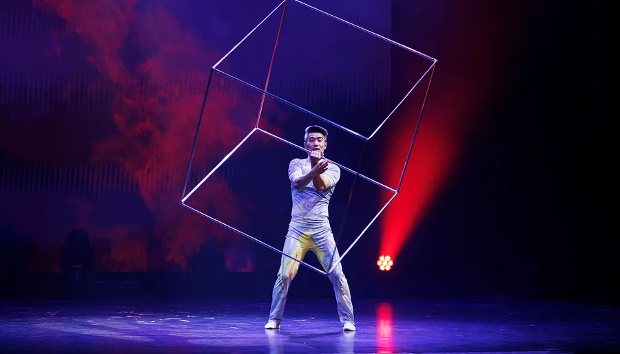 A performer manipulates a large wireframe cube on stage, surrounded by dramatic lighting.