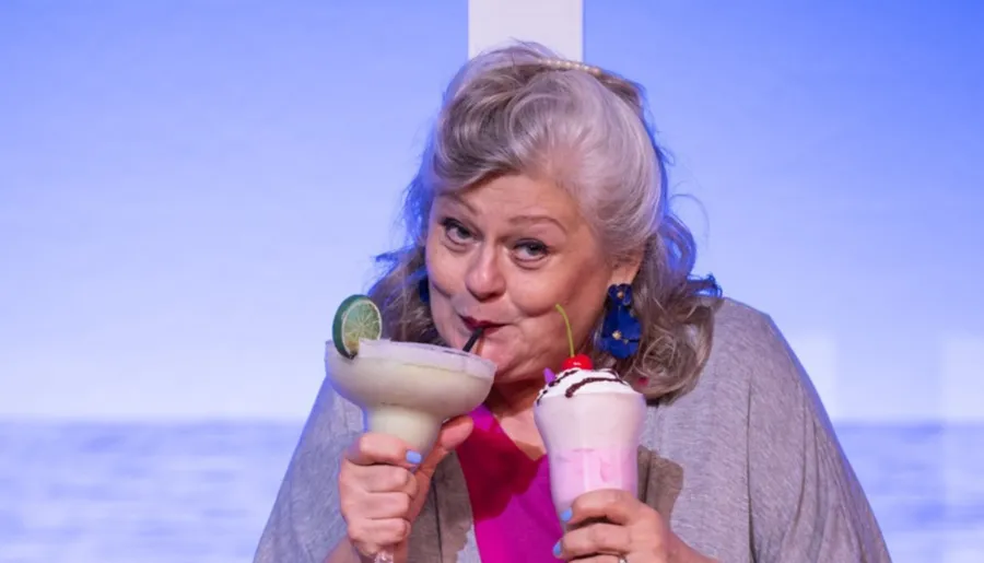 A woman is enjoying two colorful drinks, one with a lime garnish and another with whipped cream and a cherry, against a blue background.