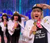 Four women wearing sailor hats and white jackets holding microphones perform enthusiastically in front of a sparkly curtain backdrop