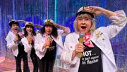Four women wearing sailor hats and white jackets, holding microphones, perform enthusiastically in front of a sparkly curtain backdrop.