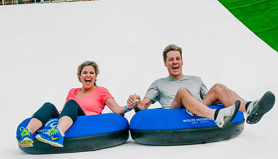 Two people are joyfully sliding down a synthetic slope on inner tubes at an outdoor recreation area.